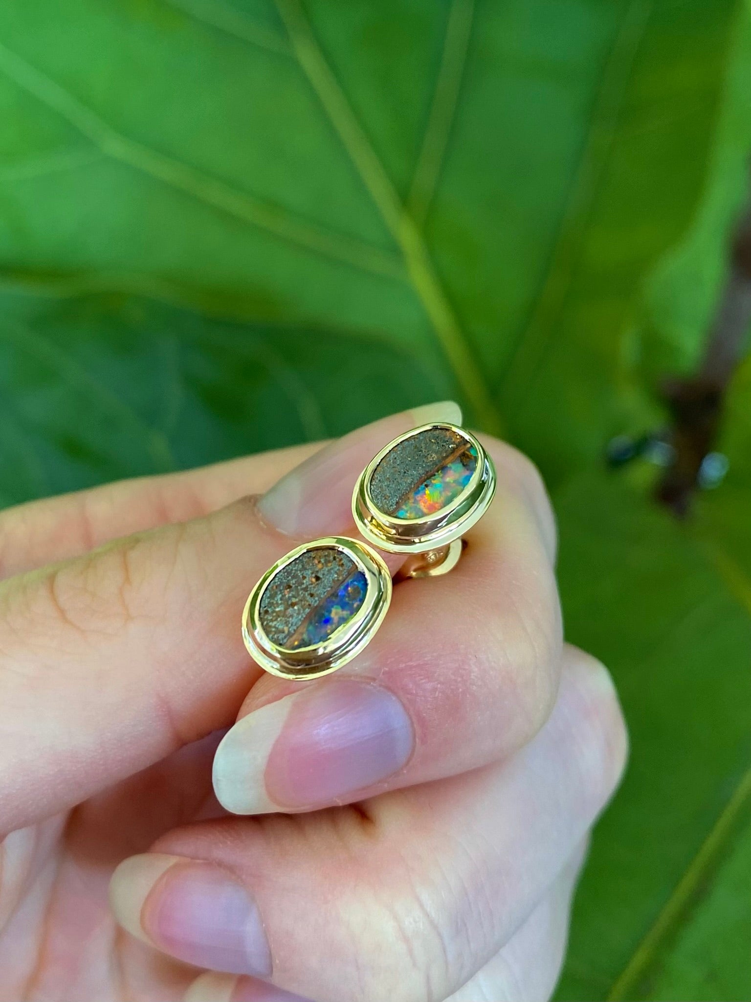 Pair of gold, bezel set earrings with opal stones held in hand against a leafy background. Opals include ironstone inclusions and a rainbow, glittery strip of opal