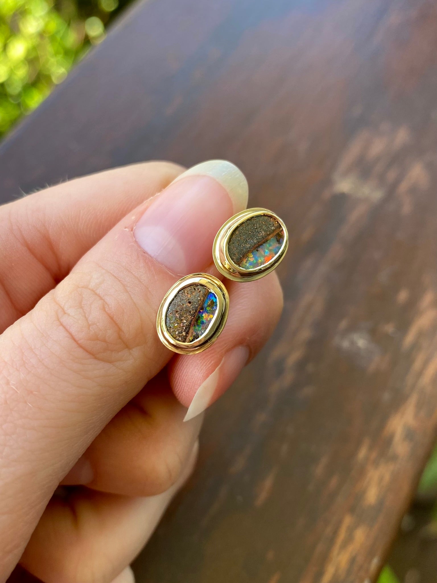 Pair of gold, bezel set earrings with opal stones hand in hand. Opals include ironstone inclusions and a rainbow, glittery strip of opal