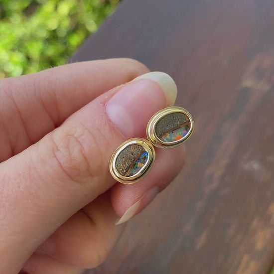 Pair of gold, bezel set earrings with opal stones held in hand. Opals include ironstone inclusions and a rainbow, glittery strip of opal