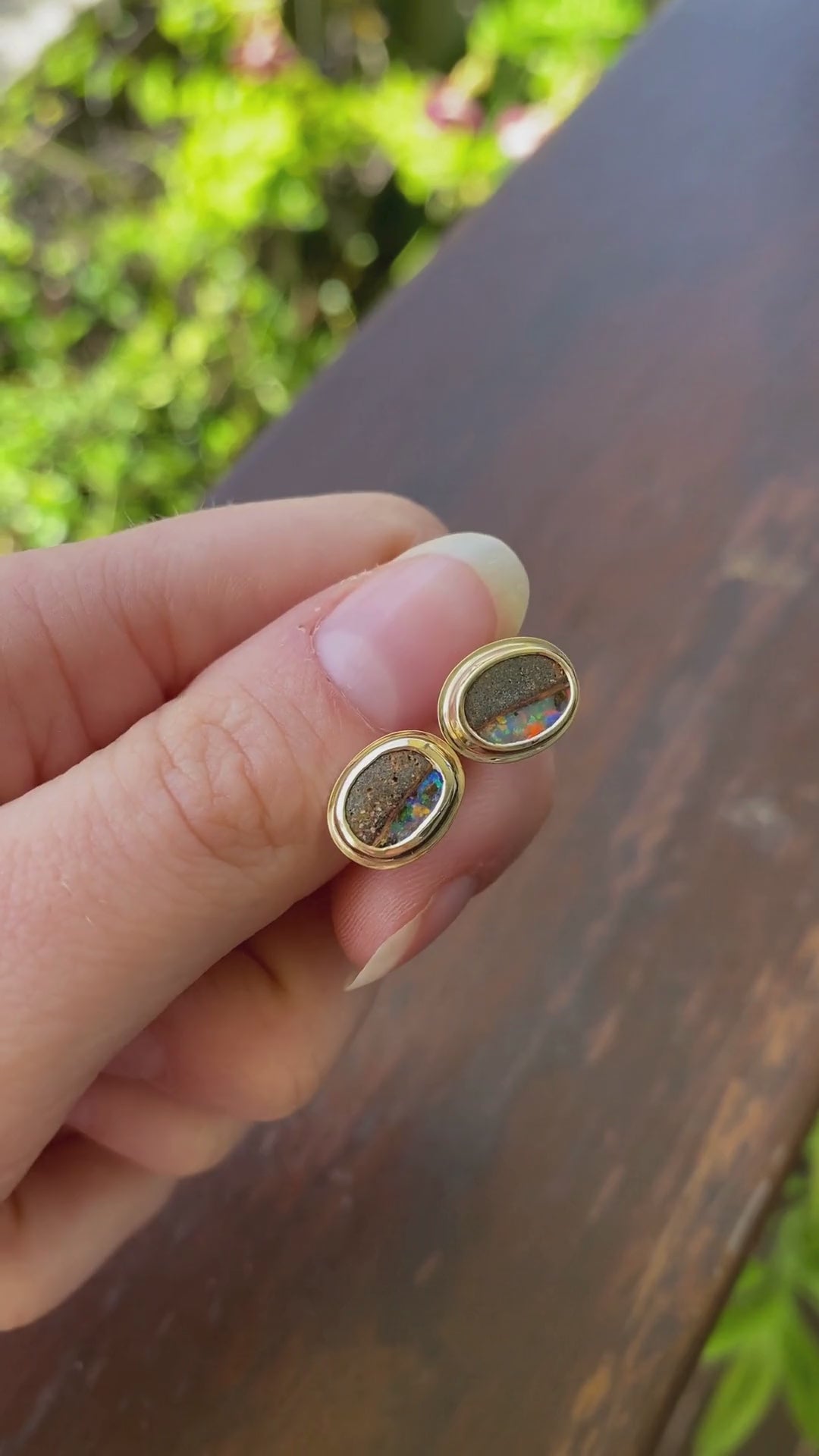 Pair of gold, bezel set earrings with opal stones held in hand. Opals include ironstone inclusions and a rainbow, glittery strip of opal