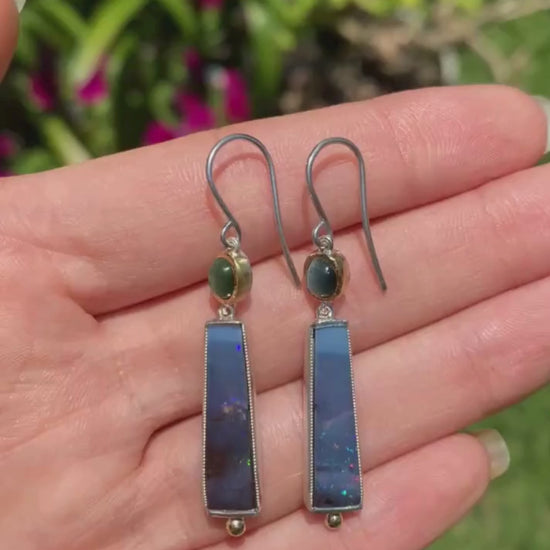 hand holding a pair of earrings with green tourmaline and blue boulder opal set in yellow gold and silver moving in the sunlight