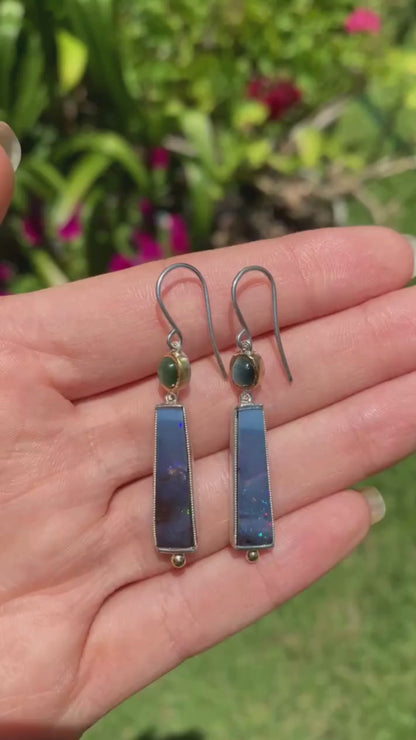 hand holding a pair of earrings with green tourmaline and blue boulder opal set in yellow gold and silver moving in the sunlight