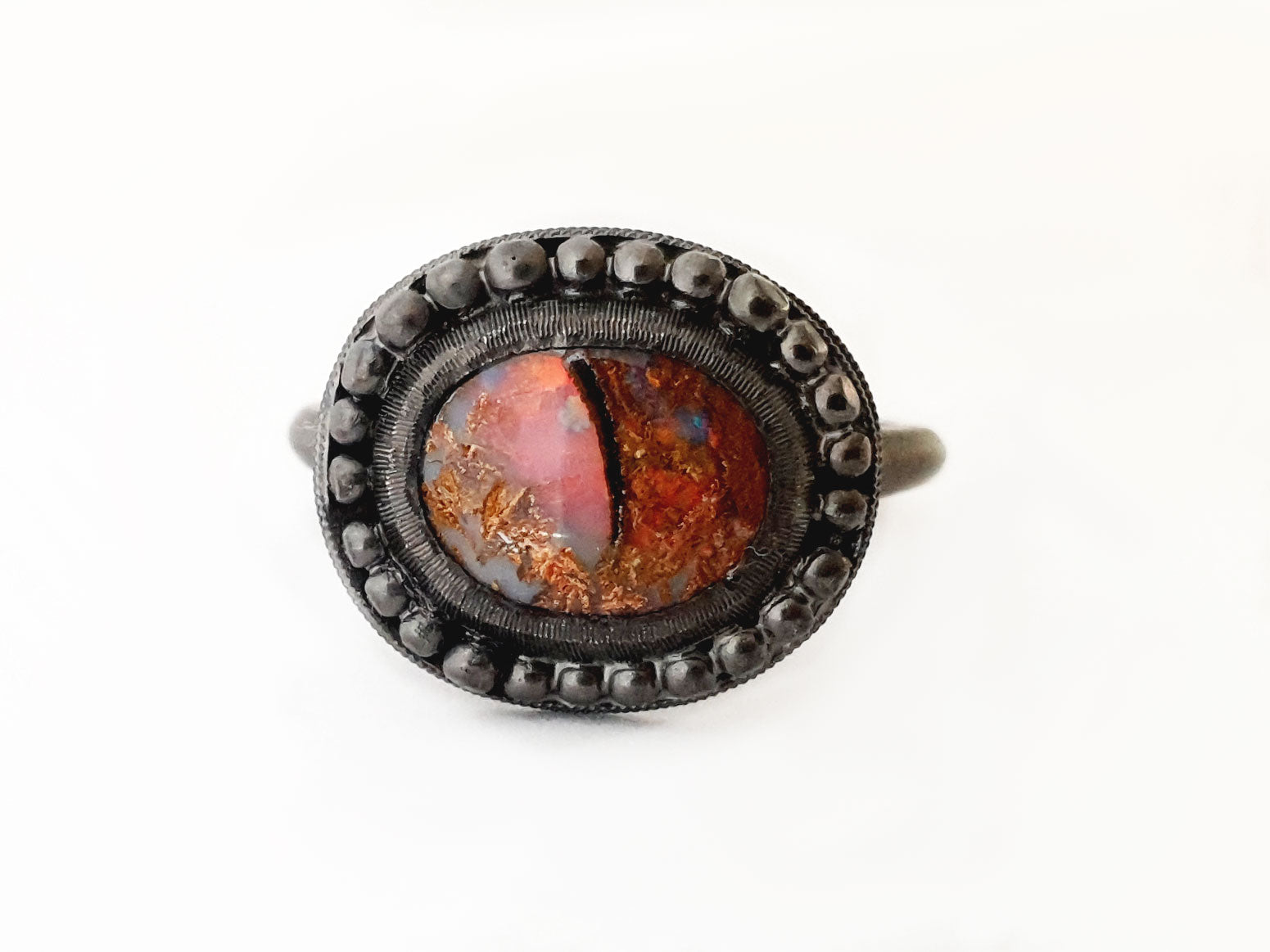 Queensland Boulder Opal ring with a blackedned silver bezel framed by silver balls. This opal looks like a dragons eye, the stone can be seen through the colours of the opal. The opal has reds blue greens and purples. Hand crafted and Australian made by Custom Jewellery Co using local and ethically sourced materials. Australian designed and made for high quality fine jewellery. 